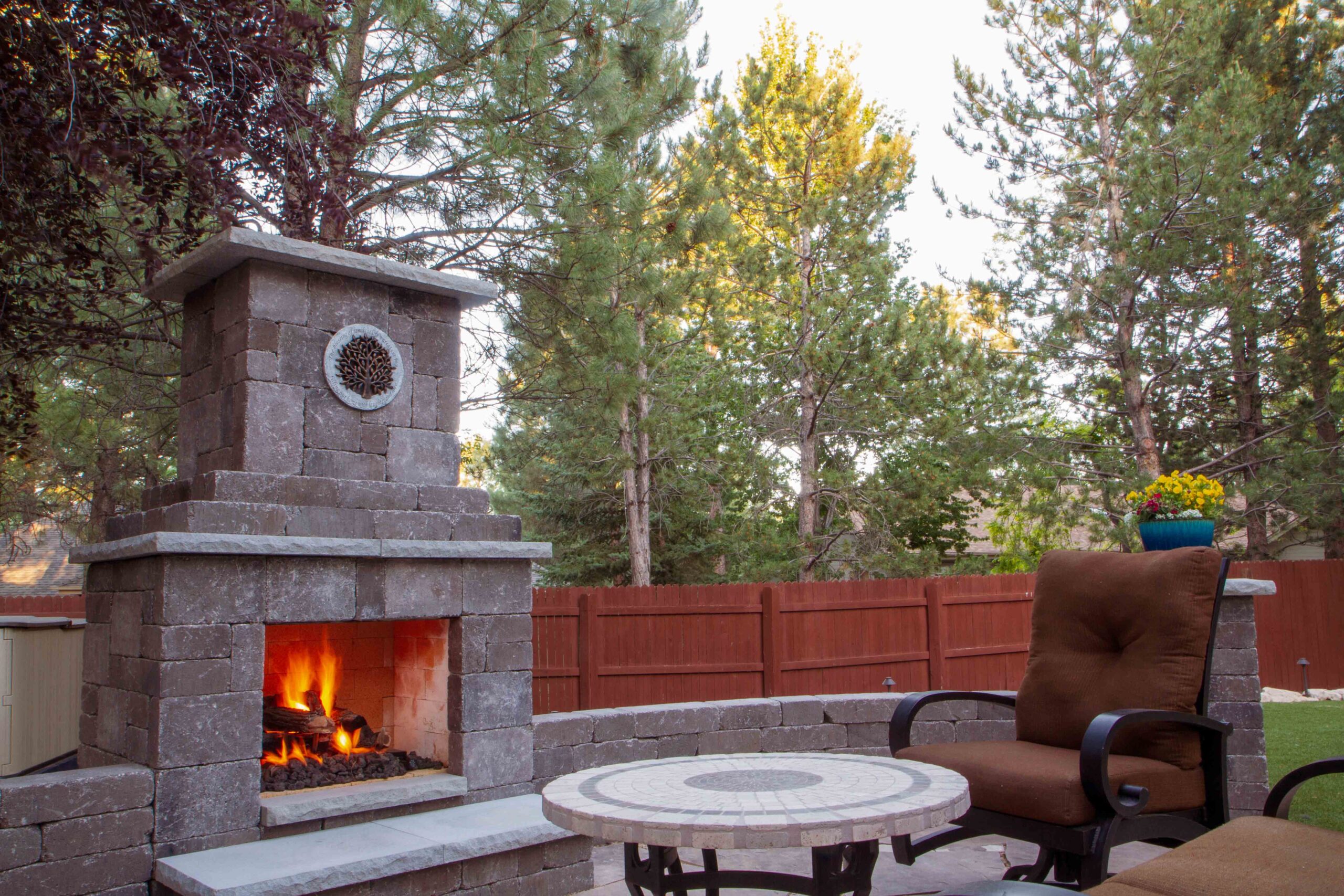 Outdoor patio with a lit fireplace and sitting area surround by pine trees